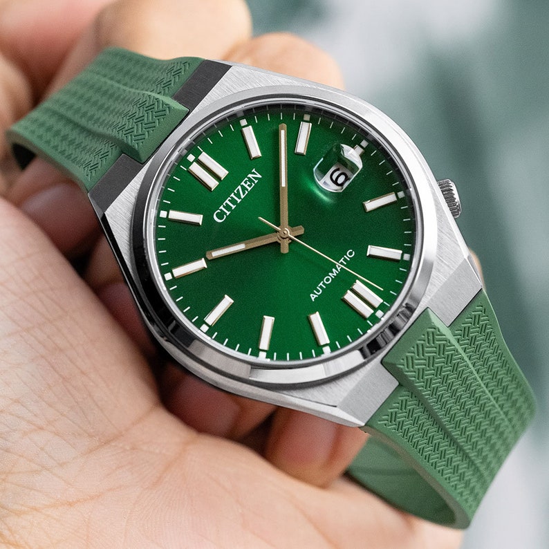Puede incluir: Un reloj autom&aacute;tico Citizen plateado con una esfera verde y una correa de caucho verde. El reloj tiene una ventana de fecha en la posici&oacute;n de las 3 en punto y marcadores de hora romanos blancos.