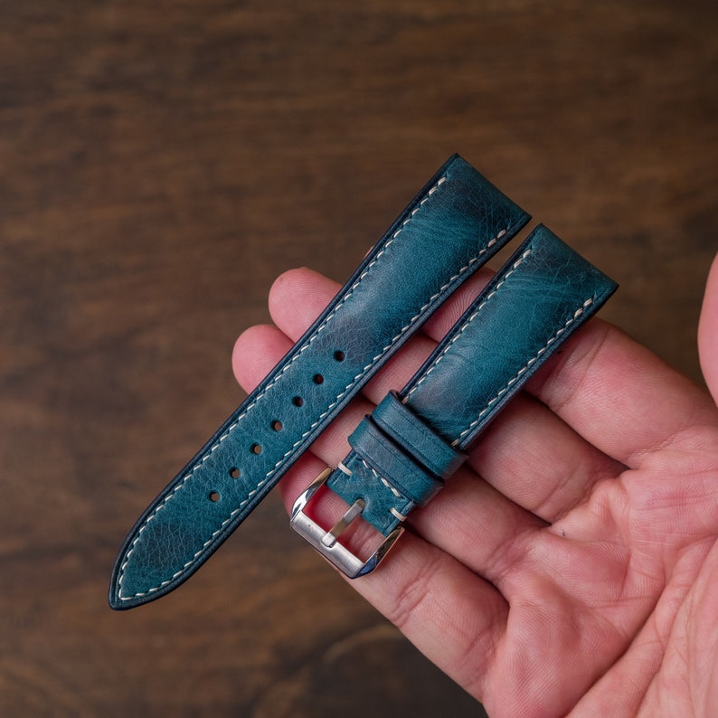 May include: Two teal blue leather watch straps with white stitching. The straps are laid out on a hand, showing the buckle and stitching details.