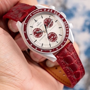May include: A silver chronograph wristwatch with a white face and red accents. The watch has a red leather strap with a crocodile pattern. The watch face features the text "OMEGA SWATCH Speedmaster MOONS WATCH".