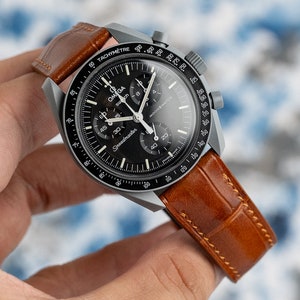 May include: A black Omega Speedmaster Moonwatch chronograph wristwatch with a brown leather strap. The watch has a black dial with white markings and a tachymeter bezel. The watch is held in a hand.