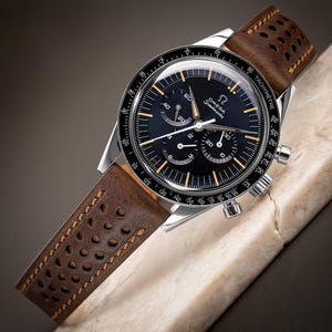 May include: A silver chronograph wristwatch with a black face and orange accents. The watch has a brown leather band with a perforated pattern.
