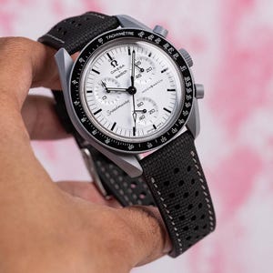 Puede incluir: Un reloj de pulsera plateado con esfera blanca y detalles en negro. El reloj presenta los logotipos de Omega y Swatch, la palabra "Speedmaster" y "MOONSWATCH". La correa negra tiene un diseño perforado.