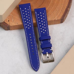 May include: Two-piece blue watch strap with white stitching and perforated holes. The strap has a textured surface and a silver-colored buckle. The watch band is displayed on a wooden surface.