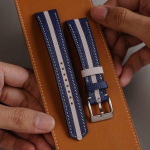 May include: Blue and white striped watch straps with white stitching. The straps have a silver buckle and are displayed on a brown leather surface. The watch straps are held by a person's hands.