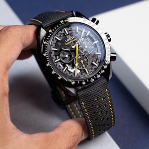 May include: A black and yellow chronograph wristwatch with a black leather strap and a tachymeter scale around the bezel. The watch face features the text "OMEGA Speedmaster" and a chronograph dial.