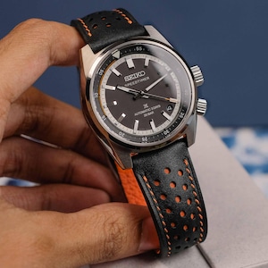 May include: A stainless steel Seiko Speedtimer watch with a black dial and a black leather strap with orange stitching. The watch face displays the text "SEIKO SPEEDTIMER AUTOMATIC 3 DAYS 20 BAR". The watch is held by a hand.