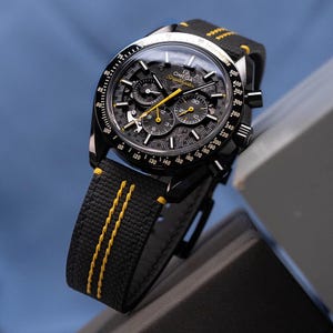 May include: A black Omega Speedmaster watch with a black dial and yellow accents. The watch features a black strap with yellow stitching. The watch face displays multiple sub-dials and the Omega Speedmaster logo.