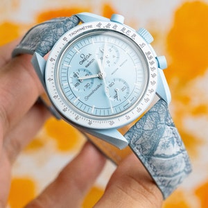 May include: A light blue Omega Speedmaster Moonwatch chronograph wristwatch with a white tachymeter scale and a gray patterned leather strap.