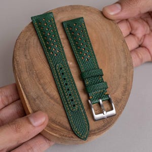 May include: A dark green watch strap featuring a textured surface, perforated detailing, and orange stitching. The strap includes a silver-toned metal buckle and two matching green leather keepers, designed for secure attachment to a timepiece.