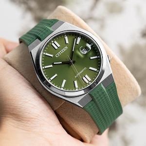 Puede incluir: Un reloj autom&aacute;tico Citizen plateado con una esfera verde y una correa de caucho verde. El reloj tiene una ventana de fecha en la posici&oacute;n de las 3 en punto y las palabras "AUTOMATIC" y "CITIZEN" impresas en la esfera.