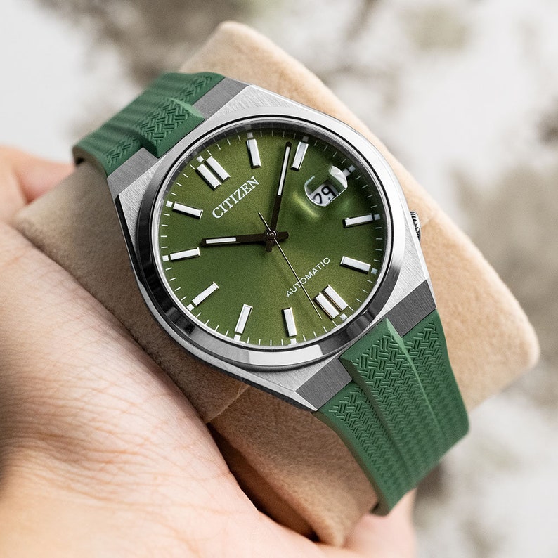 Puede incluir: Un reloj autom&aacute;tico Citizen plateado con una esfera verde y una correa de caucho verde. El reloj tiene una ventana de fecha en la posici&oacute;n de las 3 en punto y las palabras "AUTOMATIC" y "CITIZEN" impresas en la esfera.