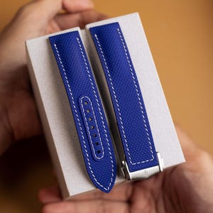 May include: A pair of royal blue watch straps featuring a textured surface and white contrast stitching. One strap includes a series of punched holes for adjustment, while the other is attached to a silver-toned metal deployment clasp buckle.