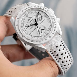 Puede incluir: Un reloj de pulsera cronógrafo Omega Speedmaster blanco con una correa de cuero blanco con un patrón perforado. La esfera del reloj tiene marcas y manecillas negras, y la escala del taquímetro está impresa en el bisel.