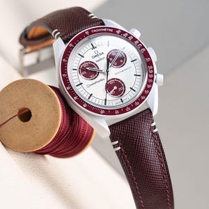 May include: A white chronograph watch with a red tachymeter bezel and a burgundy leather strap. The watch face features the text "OMEGA swatch Speedmaster" and "MOONS WATCH".