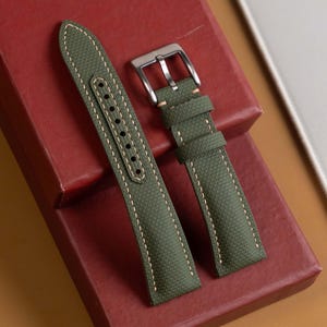 May include: A two-piece olive green watch strap featuring a textured surface, light-colored contrast stitching along the edges, and a polished silver-tone metal pin buckle. The strap includes two matching olive green loops for securing the end.