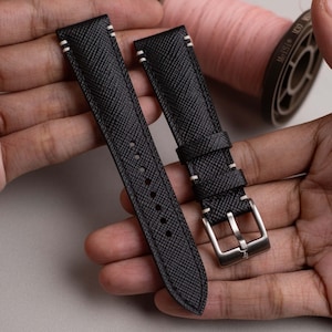 May include: A black watch strap with a cross-hatched texture, featuring white contrast stitching along the edges. The strap includes a silver-toned metal buckle and two matching leather keepers, with a series of circular adjustment holes.