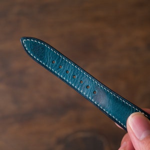 May include: A teal blue leather watch strap with white stitching and five holes for adjusting the size.