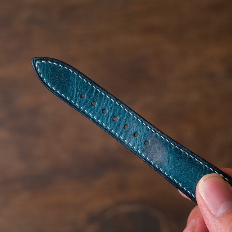 May include: A teal blue leather watch strap with white stitching and five holes for adjusting the size.