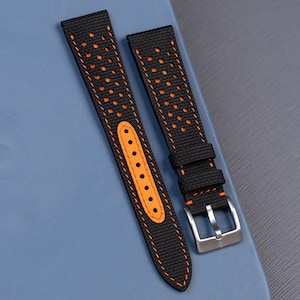 May include: A two-piece watch strap featuring a black textured surface with perforated holes revealing an orange underlayer. The strap includes orange stitching, an orange reinforced hole section, and a silver-toned metal pin buckle.