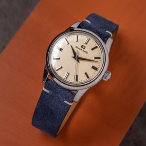 May include: A silver wristwatch with a white face and black hands. The watch has a blue suede strap and the text "Grand Seiko" on the face.