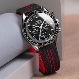 May include: A black Omega Speedmaster Professional watch with a black dial and silver case. The watch has a black strap with red stitching. The watch face includes the text "OMEGA Speedmaster PROFESSIONAL".