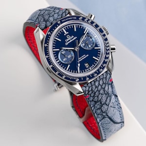 Puede incluir: Un reloj de pulsera cronógrafo plateado con una esfera azul y una correa de cuero gris y roja. La esfera del reloj tiene el texto "OMEGA Speedmaster" y "CO-AXIAL CHRONOMETER".