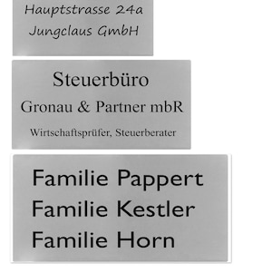 May include: A silver plaque with the text "Familie Mustermann" followed by "Jurgen & Tamara Schefer". Below that is another plaque with the text "Familie Müller". The next plaque has the text "Hauptstrasse 24a Jungclaus GmbH". The next plaque has the text "Steuerbüro Gronau & Partner mbR Wirtschaftsprüfer, Steuerberater". The final plaque has the text "Familie Pappert Familie Kestler Familie Horn".
