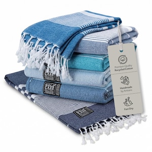 May include: A stack of five folded rectangular cotton towels in various shades of blue, featuring white striped patterns and white fringe ends. A beige rectangular tag is attached, displaying the text Premium Quality Recycled Cotton, Handmade by Artisans, and Fast Dry with corresponding icons.