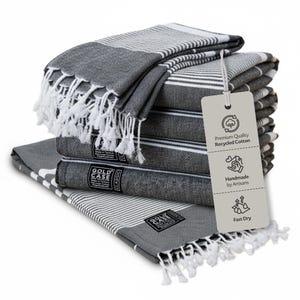 May include: A stack of charcoal gray and white striped towels with knotted fringe ends. Each towel features a black label reading GOLD CASE CLASSIC. A beige tag attached by a string displays icons and text: Premium Quality Recycled Cotton, Handmade by Artisans, and Fast Dry.