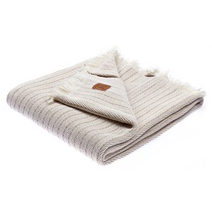 May include: A cream-colored throw blanket with a herringbone pattern and fringe edges. The blanket is folded and has a small brown leather tag with the text "The Company Store" on it.