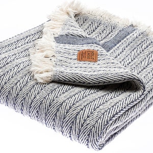 May include: A white and blue herringbone throw blanket with a fringe border. The blanket is folded and has a brown leather tag with the text "GOLD CASE" on it.