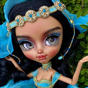 Könnte beinhalten: Eine Puppe in einem türkisfarbenen und goldenen Kostüm mit passendem Kopfschmuck. Die Puppe hat dunkles Haar, dramatisches Augen-Make-up und einen fließenden, durchsichtigen Rock. Die Puppe ist vor einem Hintergrund aus grünem Laub posiert.