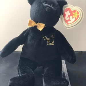 May include: Black beanie baby bear with a gold bow tie and the text "The End" embroidered on its chest. The bear is sitting on a clear plastic display stand. The bear has a Ty tag attached to its ear.