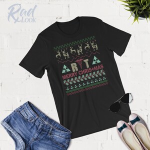 May include: Black t-shirt with a green and red Christmas-themed design. The design features a medical symbol, the letters "RTA", and the text "Merry Chris+Mas".