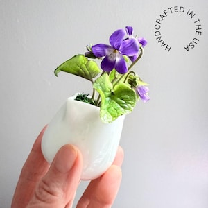 May include: A small white ceramic vase holds a miniature violet plant with purple flowers and green leaves. The vase is held in a hand. The text "HANDCRAFTED IN THE USA" is in a circle in the background.