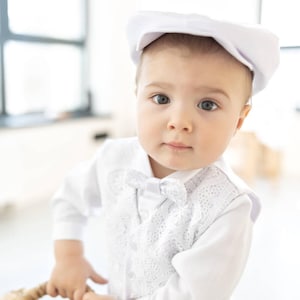 May include: A baby boy wearing a white outfit with a lace vest and a white bow tie. He is wearing a white cap and looking at the camera.