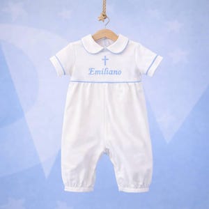 Puede incluir: Romper de bebé blanco con cuello Peter Pan y mangas cortas. El romper presenta ribetes azul claro, una cruz y el nombre "Emiliano" bordado en azul claro. La prenda está colgada en una percha de madera sobre un fondo azul claro.