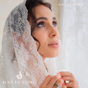 May include: A white lace veil with a delicate floral pattern. The veil is worn by a person with dark hair and is draped over their head and shoulders. The word "Battesimo" is visible in the bottom left corner of the image.