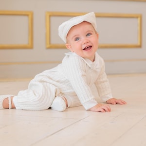May include: A cream-colored cable knit romper with a matching flat cap and soft booties. The long-sleeved one-piece garment features a collared neckline and a textured vertical cable pattern throughout, providing a coordinated outfit for an infant.