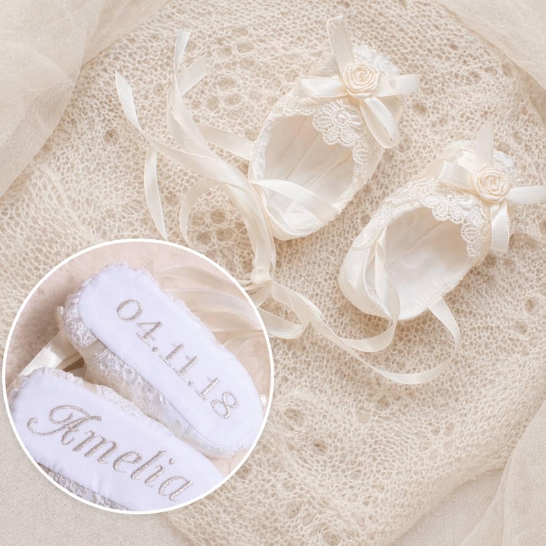 K&ouml;nnte beinhalten: Elfenbein Baby-Schuhe mit Spitzenbesatz, Satinb&auml;ndern und einer kleinen Rosenverzierung. Die Sohlen sind mit dem Namen "Amelia" und dem Datum "04.11.18" bestickt. Diese zarten Schuhe sind ideal f&uuml;r einen besonderen Anlass.