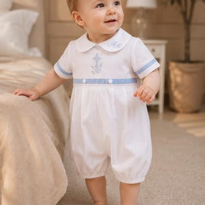 May include: A white short-sleeved romper featuring a rounded collar with embroidered initials, a light blue horizontal waistband with two buttons, and a light blue cross and dove embroidery on the chest. The garment has a gathered bubble-style bottom.