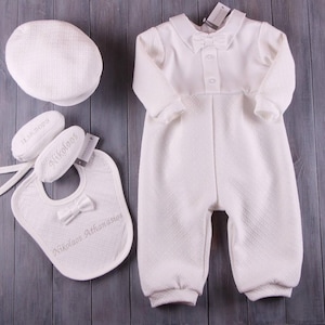 Könnte beinhalten: Ein weißes Baby-Outfit-Set, bestehend aus einem Strampler mit Fliege, einem Lätzchen, einem Hut und Babyschuhen. Das Set hat ein strukturiertes Muster und enthält den Namen "Nikolaos" und das Datum "11.18.2023" auf Lätzchen und Schuhen.