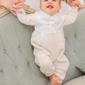 Könnte beinhalten: Ein weißer, langärmeliger Baby-Strampler mit Zopfstrickmuster an Ärmeln und Beinen, einem glatten, satinartigen Oberteil, einem spitzen Kragen und einer kleinen, passenden Fliege in der Mitte. Das Kleidungsstück hat eine Knopfleiste vorne.