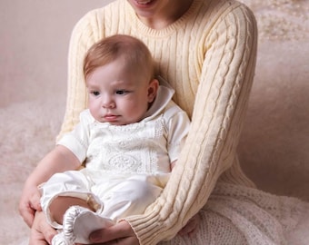Könnte beinhalten: Ein Baby in einem weißen, bestickten Strampler und passenden Babyschuhen wird von einer Person gehalten, die einen gelben Zopfstrickpullover trägt. Das Baby hat hellbraunes Haar und blickt nach unten. Der Hintergrund ist eine weiche, neutrale Farbe.