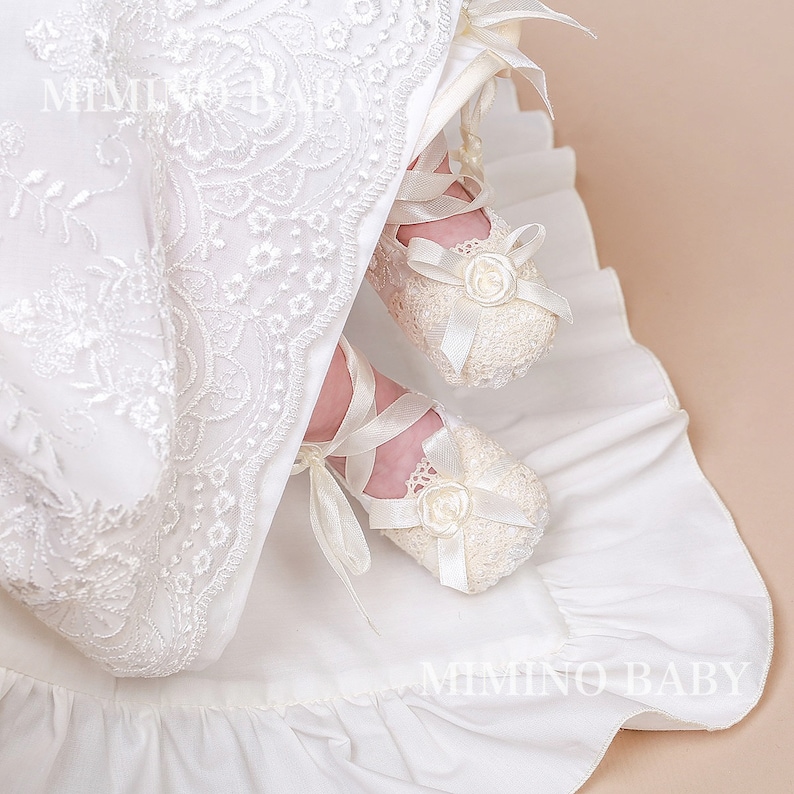 K&ouml;nnte beinhalten: Elfenbeinfarbene Baby-H&auml;kelschuhe mit Satinb&auml;ndern und einer kleinen Rosenverzierung. Die Schuhe sind Teil eines wei&szlig;en Spitzen-Taufkleides, mit dem Text "MIMINO BABY" sichtbar.