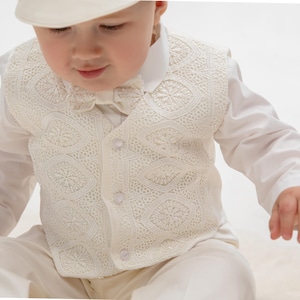 May include: A white baby vest with a delicate lace pattern and a bow tie. The vest has a button closure and is made of a soft, lightweight fabric.
