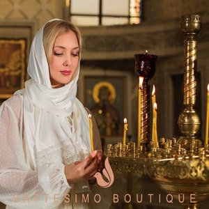 May include: A woman wearing a white headscarf stands in a church, holding a lit candle in front of a golden candle stand with many lit candles.