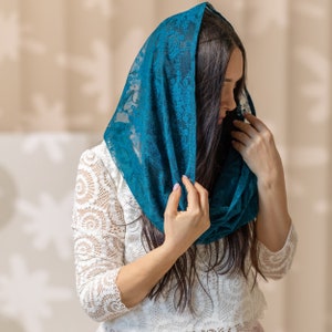 May include: A woman wearing a teal lace scarf over her head and a white lace top.