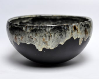 May include: A ceramic bowl with a matte black finish. The rim has a textured glaze in white, grey, and brown, creating a striking contrast. This decorative bowl is suitable for serving or display. The bowl is likely handmade.