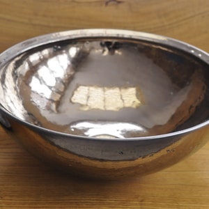K&ouml;nnte beinhalten: Eine geh&auml;mmerten Metallsch&uuml;ssel mit einem reflektierenden, silberfarbenen Innenraum. Die Sch&uuml;ssel hat eine abgerundete Form und steht auf einer Holzoberfl&auml;che. Ein zusammengerolltes graues Handtuch ist links zu sehen, was auf ein Badezimmer hindeutet.